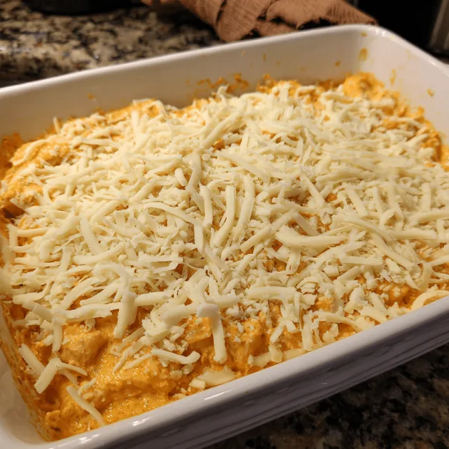 Shredded chicken and cheeses being folded into creamy Buffalo sauce mixture in mixing bowl with wooden spoon